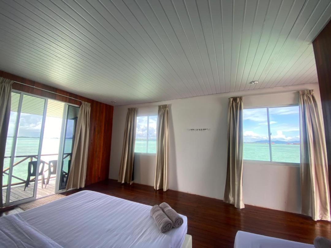 A spacious bedroom at Sea Plus Water Chalet Resort near the Tun Sakaran Marine Park providing a large bed facing floor to ceiling glass doors to the balcony, rich mahogany style floors, and AC.