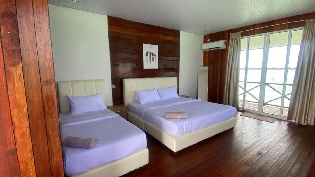 A bright bedroom at Sea Plus Water Chalet Resort near Semporna presenting two beds, glass-panel doors opening onto the private balcony, warm wooden plank floors, and air con.